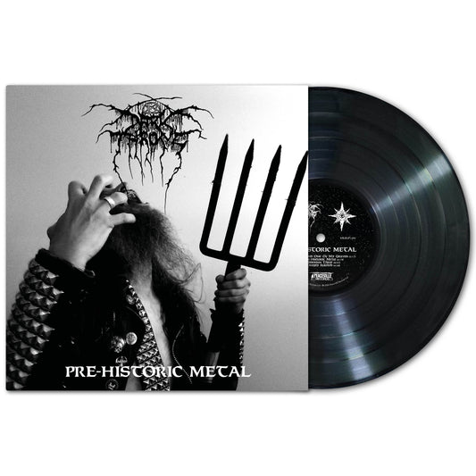 A Black vinyl record beside its cover, featuring artwork of a bearded figure in spiked clothing holding a pitchfork. The album, PRE-HISTORIC METAL by Darkthrone, has a black metal-style logo evoking classic primitive metal.